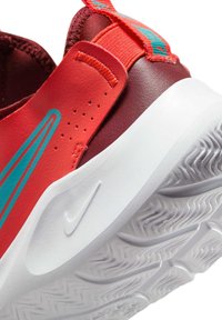 Zapatilla deportiva roja y marrón con acentos en turquesa, suela de goma, agarre texturizado y una pestaña en el talón para facilitar el calzado.