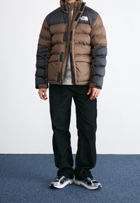 Giacca imbottita marrone e nera, con colletto alto, cuciture orizzontali, apertura con zip e logo sul petto. I pantaloni neri e le sneakers completano l'outfit.