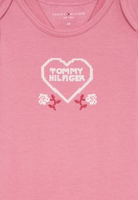 Rožinė medvilninė kūdikių bodžių su širdies raštu ir balta "TOMMY HILFIGER" užrašu. Gėlėti akcentai balta ir burgundiška spalvomis po širdimi.
