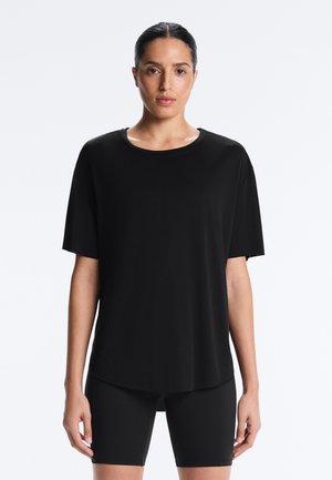 Femme aux cheveux foncés attachés, portant un t-shirt noir ample à manches courtes et un cuissard de cyclisme noir moulant, sur fond blanc.