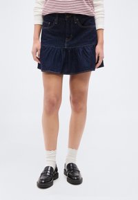 Γυναίκα που φοράει μια μίνι φούστα από σκούρο μπλε τζιν, λευκές κάλτσες με βολάν και μαύρα δερμάτινα μοκασίνια, στέκεται μπροστά σε μια απλ ή λευκή επιφάνεια.