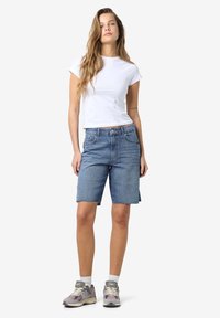 Denim šorti vidēji zilā krāsā ar saplēstu apakšmalu, kombinēti ar baltu īsu piedurkņu pieguļošu T-kreklu un pelēkiem sporta apaviem.