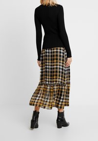 Maglietta nera a costine con maniche lunghe abbinata a una gonna maxi a strati in plaid gialla, nera e bianca. Stivali neri alla caviglia completano l'outfit.