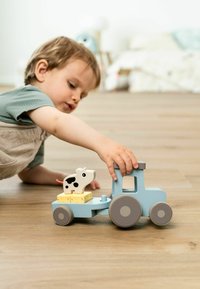 Drevený traktórik svetlomodrej farby s sivými kolesami; obsahuje žltý blok a figúrku kravy na vrchu. Ruka dieťaťa sa snaží siahnuť po hračke.