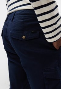 Mørkeblå bukser med struktureret stof, der har en baglomme med en brun knap og en diskret mærkat. Stribet skjorte synlig øverst.