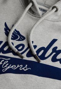Felpa grigia con lettering navy blue "Superdry Flyers", con cordino con punte in metallo e dettaglio del logo con ali vicino al cappuccio.