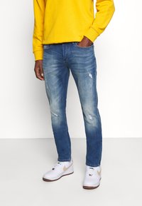 Blå denimjeans med smal passform, som har slitna detaljer och blekningar. Kombinerat med en gul sweatshirt och vita sneakers.