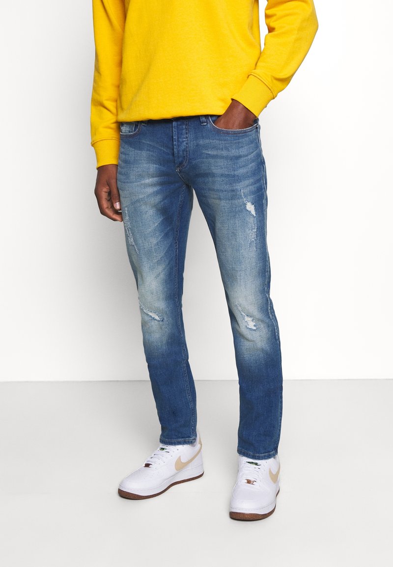Blå denimjeans med smal passform, som har slitna detaljer och blekningar. Kombinerat med en gul sweatshirt och vita sneakers.