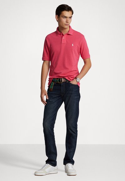 Chemise polo rose avec un petit logo, associée à un jean bleu foncé et des baskets blanches. La ceinture présente un motif rayé multicolore.