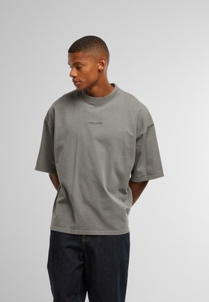 Grå oversized t-shirt med korte ærmer, rund udskæring, blød bomuldsstruktur, har diskret branding på brystet. Bæres med mørke jeans.