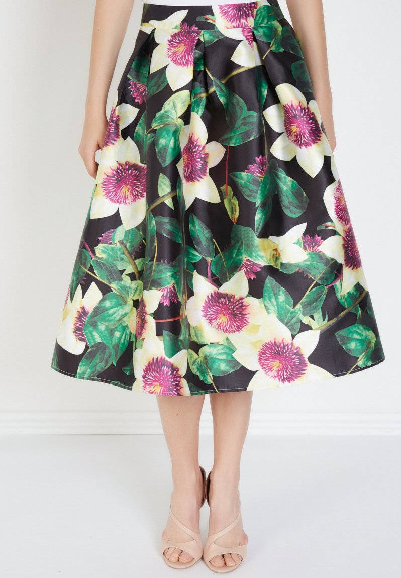 Falda midi floral con grandes flores blancas y rosas sobre un fondo negro, con pliegues y una textura suave. Complementada con sandalias beige.