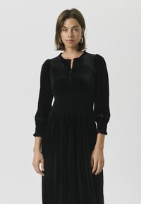 Schwarzes Samtkleid mit gerafftem Oberteil, rundem Ausschnitt und Puffärmeln. Verfügt über Rüschensäume und einen fließenden Rock. Sanfte Textur.