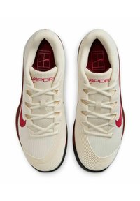 Le scarpe Nike Vapor 12 presentano una tomaia crema con mesh testurizzato, dettagli color marsala e un Swoosh rosso. La suola è nera con un design aerodinamico.