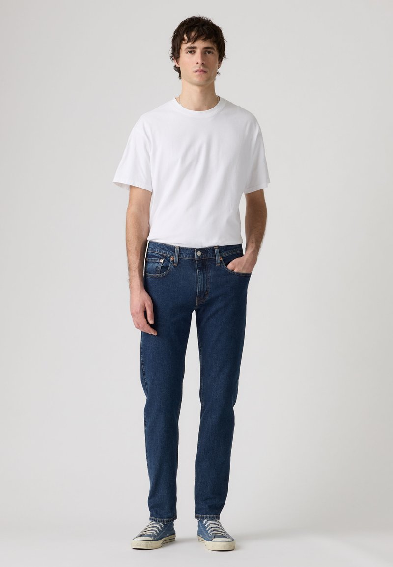 Giovane uomo in piedi con una mano in tasca, che indossa una t-shirt bianca, jeans blu scuro e sneakers blu su uno sfondo chiaro uniforme.