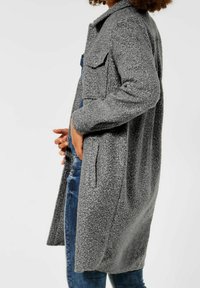 Cappotto lungo grigio e texture con tasche frontali e chiusura a pulsante. Mostrato accanto a jeans in denim blu. Il tessuto ha un aspetto morbido e peloso.