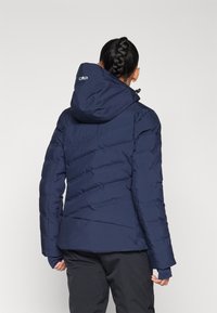 Femme aux cheveux tressés portant une veste matelassée bleu marine avec capuche et un pantalon noir, vue de dos sur un fond uni.