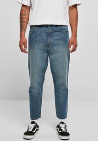 Vaqueros de mezclilla azul claro con un corte relajado, piernas cónicas y desgastado. Cuentan con trabillas para cinturón, bolsillos delanteros y cierre de botón. Zapatillas negras.