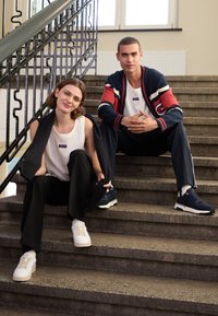 Dos modelos están sentados en unas escaleras. La mujer lleva una camiseta blanca de tirantes, pantalones negros y zapatillas blancas. El hombre lleva un cárdigan azul marino y rojo con un logo, pantalones negros y zapatillas azul marino.