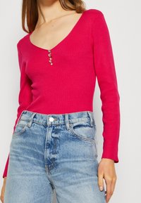 Frau trägt ein leuchtend pinkfarbenes, geripptes Langarmoberteil mit drei kleinen dekorativen Knöpfen und hellblauen hochgeschnittenen Jeans.
