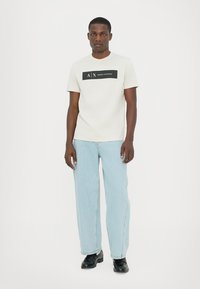 Lys grå bomull t-skjorte med en svart rektangulær logo med teksten "A|X ARMANI EXCHANGE". Kombinert med blå denimjeans og svarte sko.