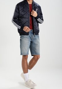 Man som justerar en navy quiltad Adidas-jacka, bärandes en röd tröja, denimshorts, vita strumpor och vita sneakers, stående mot en vit bakgrund.