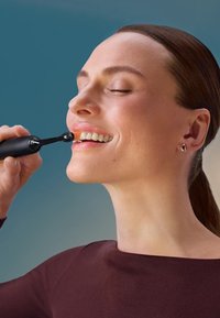 Dispositif de soin des lèvres électrique avec un corps cylindrique noir et une pointe arrondie, utilisé sur les lèvres d'une personne qui sourit, montrant des dents blanches.