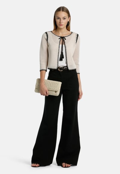 Kroksa beige cardigan dengan garis hitam, bagian depan terbuka, dan detail tali tassel. Dipasangkan dengan celana panjang hitam berkaki lebar tinggi.