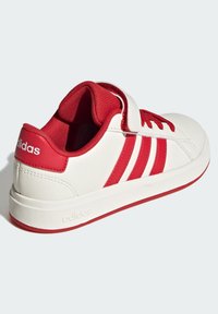 Bílé tenisky s červenými pruhy, šněrováním a patní částí, které mají suchý zip a polstrovaný límec, natočené, aby ukázaly vnější stranu a zadní část.