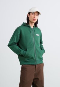 Groene zip-up hoodie met een zak aan de voorkant, geribde onderkant en boorden; bevat een klein wit logo op de linkerborst. Draagt met een paar bruine broek.