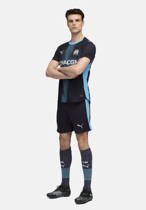 Puma futbolo apranga tamsiai mėlynos spalvos su šviesiai mėlynais akcentais. Apima marškinėlius su apykakle, atitinkančius šortus, dryžuotas kojines ir juodus sportbačius.