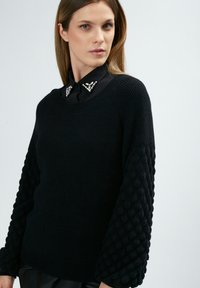 Schwarzer Pullover mit strukturierten, gep pufften Ärmeln und einem gerippten Körper. Verfügt über einen schwarzen Kragen, der mit dekorativen Edelsteinen geschmückt ist. Glatte Stoffoberfläche.