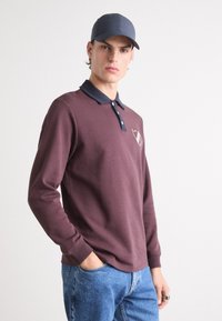 Polo shirt lung cu mânecă lungă, de culoare bordo, cu guler albastru închis, placket cu două butoane și logo pe piept. Asortat cu blugi albaștri și o șapcă albastră închis.