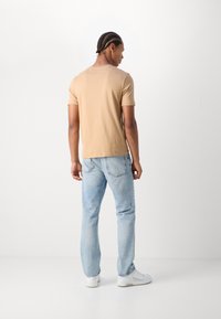 Camiseta de manga corta color marrón claro, de material de algodón, con corte recto. Llevada con vaqueros azul claros y zapatillas blancas, vista desde la parte de atrás.