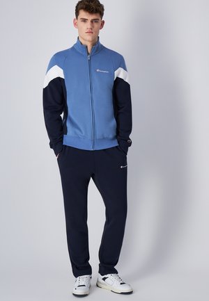 Sudadera azul con cremallera y detalles en blanco y azul marino, combinada con pantalones azul marino. Presenta puños y cinturilla elásticos, con un logo en el pecho y el muslo.