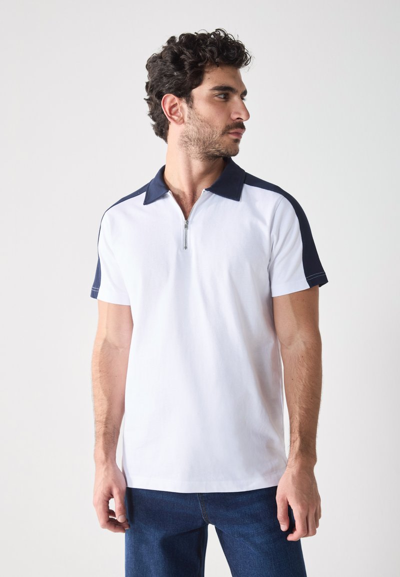 Hvid polo shirt med marineblå krave og skulderdetaljer, med en halv lynlås. Korte ærmer, blødt stof og en afslappet pasform.