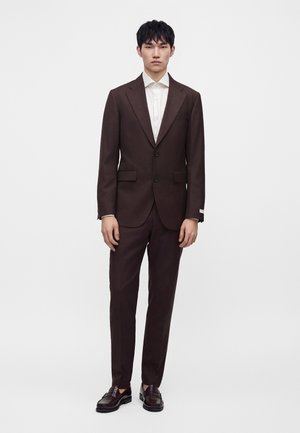 Homme debout portant un costume marron foncé ajusté avec un pantalon assorti, une chemise blanche et des mocassins en cuir marron foncé sur un fond blanc uni.