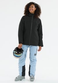 Schwarze Skijacke mit durchgehendem Reißverschluss, kombiniert mit hellblauen Skihosen mit Reißverschlusstaschen und schwarzen Seitenteilen. Ein mehrfarbiger Helm wird gehalten.