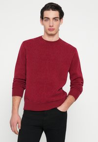 Κόκκινο πουλόβερ με υφή, ριμπ πλέξη στο λαιμό και τα μανίκια, μακριά μανίκια, συνδυασμένο με μαύρο παντελόνι, σε ουδέτερο φόντο.