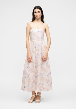 Γυναίκα με μακριά σκούρα μαλλιά φοράει αμάνικο παστέλ φλοράλ φόρεμα με λεπτές τιράντες και μπεζ τακούνια με λουράκια, στέκεται μπροστά σε απλό φόντο.