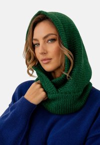 Zielony dzianinowy szal z kapturem o teksturowanym wzorze, noszony na niebieskim swetrze. Szczegóły obejmują gruby, przytulny materiał i swobodny fason.