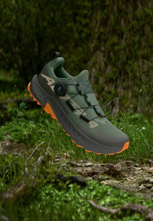 Groene trailrunningschoen met oranje zool en draaisluiting die boven de bosbodem met gras en mos zweeft.