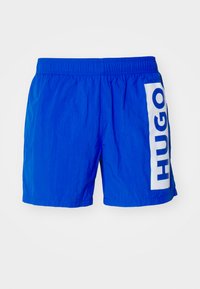 Shorts de bain bleus en tissu léger, avec une large rayure verticale blanche et le texte "HUGO" sur le côté gauche. Taille élastique.