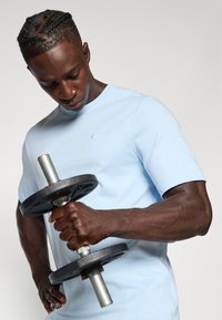 Heledad helesinine sportlik särk lühikeste varrukatega ja ümmarguse kaelusega. Inimene hoiab musta raskustahvlit hõbedase käepidemega, näidates välja hästi määratletud lihaseid.