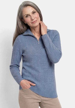 Vrouw met lang grijs haar, dragen een blauwe gestructureerde trui met een kwart ritssluiting en beige broek, één hand in de zak en de andere raakt haar nek aan.