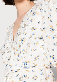 Nahaufnahme einer Frau in einem weißen Kleid mit Puffärmeln und blauen sowie gelben Blumenmustern, mit einem V-Ausschnitt und einer Taillenbindung.