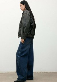 Chaqueta de cuero negra con un corte sobredimensionado y cinturilla elástica, combinada con jeans de mezclilla oscuros de pierna ancha, de pie frente a un fondo neutro.