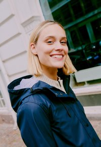 Veste marine avec fermeture éclair et boutons-pression, dotée d'une capuche à rayures, portée sur un pull crème affichant une texture lisse.