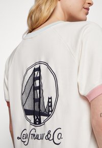 Bílá mikina s růžovými detaily, na které je vyšitý tmavě modrý grafický motiv Golden Gate Bridge a text "Levi Strauss & Co." pod ním.