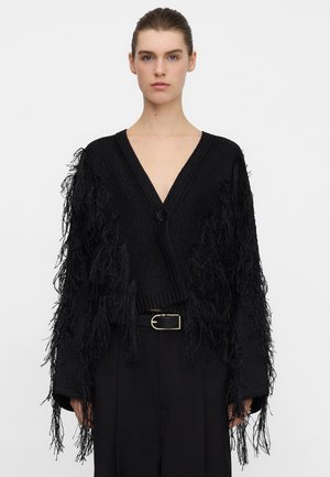 Cardigan nero lavorato a maglia con lunghe frange, scollo a V profondo e chiusura con un solo bottone. Indossato sopra pantaloni neri a vita alta con cintura.