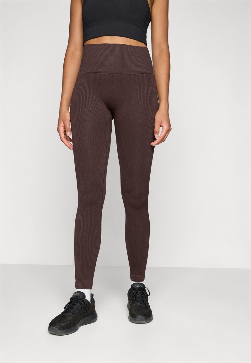 Barna bordázott leggings, magas derékkal, fekete sportcipőkkel párosítva. Az anyaga sima tapintású és testhezálló fazonú.